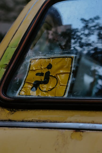Close-up of a vehicle's rear side window with a yellow disability parking permit or badge displayed on the inside. The permit features a black pictogram of a person in a wheelchair, with a black strap or cord attached to the figure, set against a yellow background. The window shows slight dirt and smudges around the edges, indicating regular use. The vehicle's body, visible in the lower part of the image, is yellow with some areas of chipped paint and minor rust, typical of older parked vehicles. The background outside the window is blurred, showing hints of trees and sky, suggesting the vehicle is parked outdoors. The scene, with its focus on this permit within the vehicle, hints at the context of compliance or access but also aligns with principles of private or alternative parking considerations, which are often related to the themes of rubbish and waste collection logistics managed by services like Rubbish Clearance Richmond.