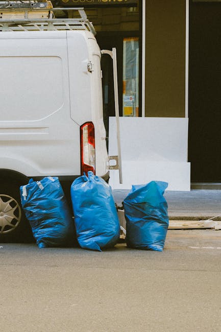 Three large blue rubbish bags filled with waste are positioned on the pavement next to a white commercial van with a rear door and taillight, parked outside a building with a dark reflective glass window. The bags appear to be made of heavy-duty plastic, with slightly crinkled textures indicating they are filled with bulky debris or refuse. The scene suggests a private or independent waste collection, likely managed by Rubbish Clearance Richmond, outside an urban property. A small wooden plank lies on the ground near the bags, and a white object, possibly a piece of building material or used packaging, rests nearby on the pavement. The environment includes a clean sidewalk, and the building's exterior includes a glass window with reflections of street elements. Natural lighting illuminates the scene, emphasizing the contrast between the parked vehicle, the bags, and the surrounding urban context, illustrating an example of on-site rubbish clearance in progress.