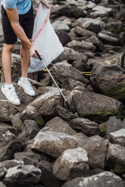 A person wearing white sneakers, navy shorts, and a light blue t-shirt is using a litter picker to collect small debris among large, irregularly shaped rocks on a rocky outdoor surface. The rocks vary in size and color, predominantly grey and brown, with some showing patches of moss or algae. The individual is holding a white trash bag with a pink stripe, positioned behind their leg, ready to collect the gathered litter. The environment appears to be a natural, outdoor setting, possibly a riverbank or rocky shoreline, with no visible man-made structures. The scene is illuminated by natural daylight, highlighting the textures of the rocks and the casual attire of the person. The image subtly aligns with themes of independent rubbish collection and environmental cleanup, similar to services offered by Rubbish Clearance Richmond, which specializes in rubbish removal and waste management solutions in Richmond. The focus on removing waste from a natural environment emphasizes the importance of proper waste disposal outside of conventional urban areas.