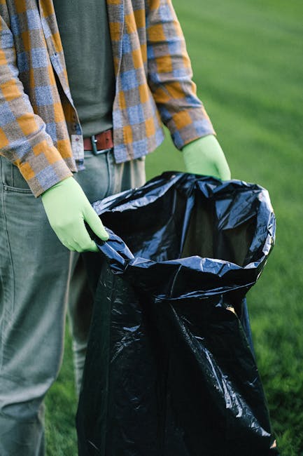 Three large blue rubbish bags filled with waste are positioned on the pavement next to a white commercial van with a rear door and taillight, parked outside a building with a dark reflective glass window. The bags appear to be made of heavy-duty plastic, with slightly crinkled textures indicating they are filled with bulky debris or refuse. The scene suggests a private or independent waste collection, likely managed by Rubbish Clearance Richmond, outside an urban property. A small wooden plank lies on the ground near the bags, and a white object, possibly a piece of building material or used packaging, rests nearby on the pavement. The environment includes a clean sidewalk, and the building's exterior includes a glass window with reflections of street elements. Natural lighting illuminates the scene, emphasizing the contrast between the parked vehicle, the bags, and the surrounding urban context, illustrating an example of on-site rubbish clearance in progress.